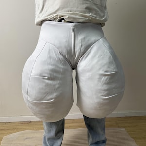 May include: A pair of white, voluminous, balloon-shaped trousers featuring a high-waisted design with a central vertical zipper closure. The garment has a smooth, matte fabric texture and exaggerated, rounded hip sections that taper down toward the legs.