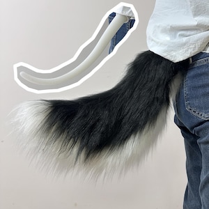 May include: A long, fluffy faux fur tail featuring a black and white colour pattern. The tail is attached to a white belt or waistband. A separate, smooth, white, curved, rigid tail-shaped structure is shown above with a white outline.