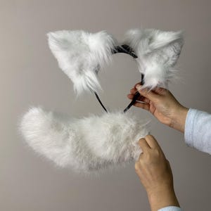 Op de afbeelding: Een witte faux fur kattenoren hoofdband en staart set. De oren zijn bevestigd aan een zwarte hoofdband en de pluizige staart is apart. De set is ontworpen voor cosplay of kostuumgebruik.