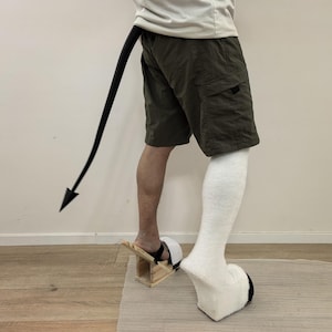 May include: A long black pointed tail attached to the waist of olive green cargo shorts. One foot wears a wooden platform sandal with a black strap, while the other foot is covered in a white fuzzy sock with a black hoof-like platform base.