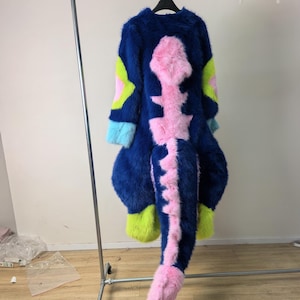 Può includere: Un costume integrale in pelliccia sintetica con una base blu scuro e un design prominente della colonna vertebrale e della coda in rosa. L'indumento presenta accenti verde neon sulle maniche e sulle sezioni inferiori, con polsini azzurri.
