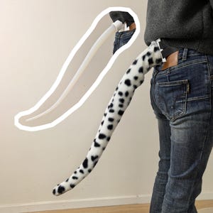 May include: A long, plush, white tail with black spots attached to a belt. The tail features a soft, faux fur texture and is worn at the waist. A white outline drawing of the tail is visible in the upper left corner of the image.