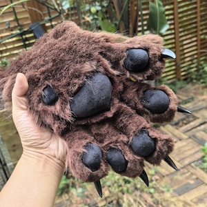 May include: Brown faux fur bear paw gloves with black paw pads and black claws. The gloves are designed to resemble a bear's paw, with a textured fur appearance and detailed paw pads.