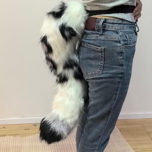 May include: A white faux fur tail with black markings, attached to a person's waist. The tail is long and fluffy, with a rounded tip. The person is wearing blue jeans.