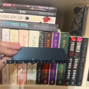 K&ouml;nnte beinhalten: Ein dunkelblaugr&uuml;ner Buchst&auml;nder mit dem ausgeschnittenen Wort "DEVOUR", der vor einem B&uuml;cherregal steht. Die B&uuml;cher haben verschiedene Farben und Titel, darunter "Birthday Girl" und "Vile Boys".