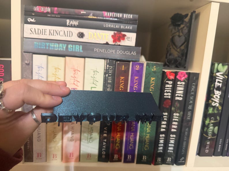 K&ouml;nnte beinhalten: Ein dunkelblaugr&uuml;ner Buchst&auml;nder mit dem ausgeschnittenen Wort "DEVOUR", der vor einem B&uuml;cherregal steht. Die B&uuml;cher haben verschiedene Farben und Titel, darunter "Birthday Girl" und "Vile Boys".