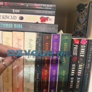 K&ouml;nnte beinhalten: Ein blau glitzerndes Wortkunstwerk mit der Aufschrift "DEVOUR ME" wird vor einem B&uuml;cherregal mit B&uuml;chern gehalten. Die B&uuml;cher haben farbige Buchr&uuml;cken und Titel. Ein dekoratives schwarzes St&uuml;ck mit Rosen befindet sich auf dem Regal.