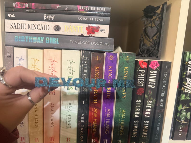 K&ouml;nnte beinhalten: Ein blau glitzerndes Wortkunstwerk mit der Aufschrift "DEVOUR ME" wird vor einem B&uuml;cherregal mit B&uuml;chern gehalten. Die B&uuml;cher haben farbige Buchr&uuml;cken und Titel. Ein dekoratives schwarzes St&uuml;ck mit Rosen befindet sich auf dem Regal.