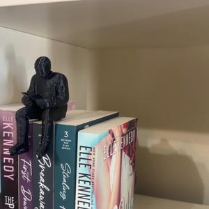 May include: A black figurine of a person reading, perched on top of a stack of books. The books have colorful spines with titles like "Kennedy" and "Stealing". The figurine is detailed and adds a decorative touch to the bookshelf.