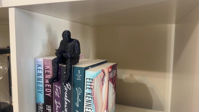 May include: A black figurine of a person reading, perched on top of a stack of books. The books have colorful spines with titles like "Kennedy" and "Stealing". The figurine is detailed and adds a decorative touch to the bookshelf.