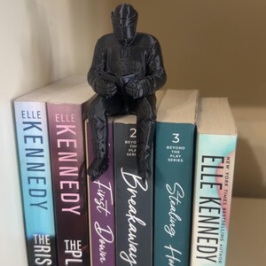 May include: A black figurine of a person sitting on a stack of books. The books have titles like "The Risk", "The Play", "Breakaway", and "Stealing Home". The books are on a shelf.