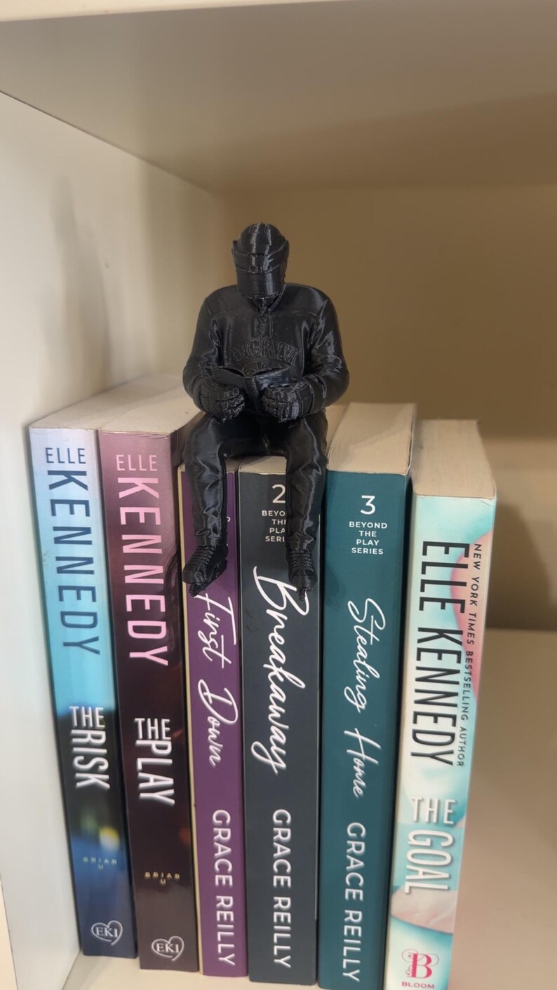 May include: A black figurine of a person sitting on a stack of books. The books have titles like "The Risk", "The Play", "Breakaway", and "Stealing Home". The books are on a shelf.