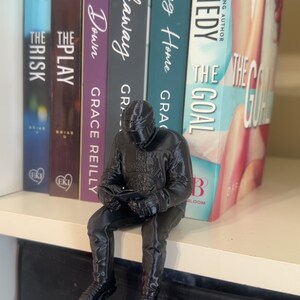 May include: A black 3D-printed figurine of a person sitting and reading, placed in front of a row of books. The books have titles like "The Goal" and "Breakaway". The figurine is detailed, with visible clothing and a helmet.