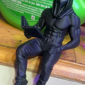Puede incluir: Una figura negra impresa en 3D de una figura musculosa con una m&aacute;scara de Scream y leyendo un libro. La figura est&aacute; sentada, con pantalones y zapatos. La figura est&aacute; sobre una superficie de madera.
