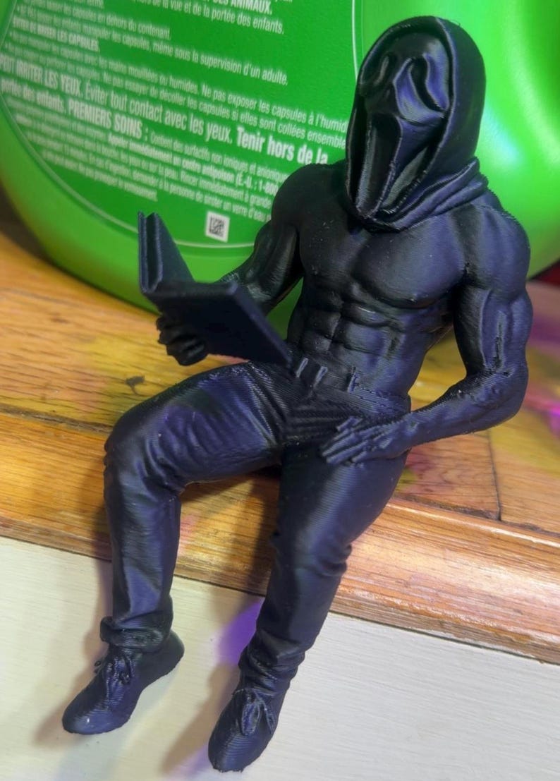 Puede incluir: Una figura negra impresa en 3D de una figura musculosa con una m&aacute;scara de Scream y leyendo un libro. La figura est&aacute; sentada, con pantalones y zapatos. La figura est&aacute; sobre una superficie de madera.