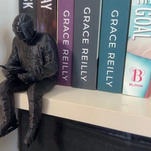 May include: A black figurine of a person in a helmet and suit, seated and reading a book, perched on a white shelf. The figurine is next to a row of books with titles including "Grace Reilly" and "The Goal."