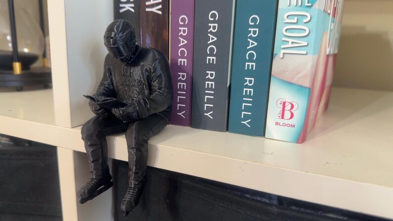 May include: A black figurine of a person in a helmet and suit, seated and reading a book, perched on a white shelf. The figurine is next to a row of books with titles including "Grace Reilly" and "The Goal."