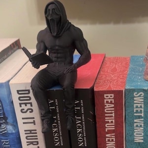 Puede incluir: Una figura negra de un personaje musculoso con una m&aacute;scara de Scream leyendo un libro, posada sobre una pila de libros. Los libros tienen t&iacute;tulos como "Does It Hurt" y "Sweet Venom". Se ve una calavera decorativa.