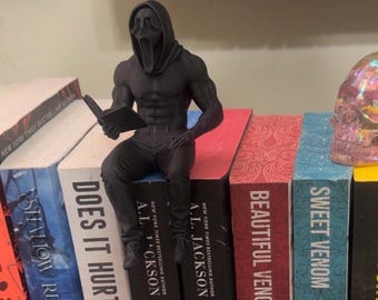 Puede incluir: Una figura negra de un personaje musculoso con una máscara de Scream leyendo un libro, posada sobre una pila de libros. Los libros tienen títulos como "Does It Hurt" y "Sweet Venom". Se ve una calavera decorativa.