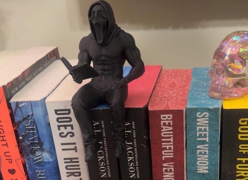 Puede incluir: Una figura negra de un personaje musculoso con una m&aacute;scara de Scream leyendo un libro, posada sobre una pila de libros. Los libros tienen t&iacute;tulos como "Does It Hurt" y "Sweet Venom". Se ve una calavera decorativa.
