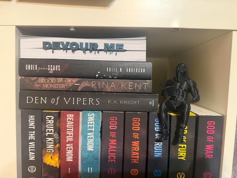 K&ouml;nnte beinhalten: Ein B&uuml;cherregal mit B&uuml;chern, darunter Titel wie "Devour Me", "Under Your Scars" und "Den of Vipers". Eine schwarze Figur eines Kriegers sitzt auf den B&uuml;chern. Die Buchr&uuml;cken sind in verschiedenen Farben gehalten.
