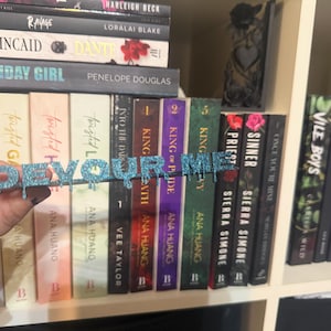 K&ouml;nnte beinhalten: Ein glitzerndes blaues "DEVOUR ME" Buchzubeh&ouml;r wird vor einem B&uuml;cherregal mit B&uuml;chern gehalten. Der Text hat einen tropfenden Effekt. Die B&uuml;cher haben farbenfrohe Buchr&uuml;cken und verschiedene Titel.