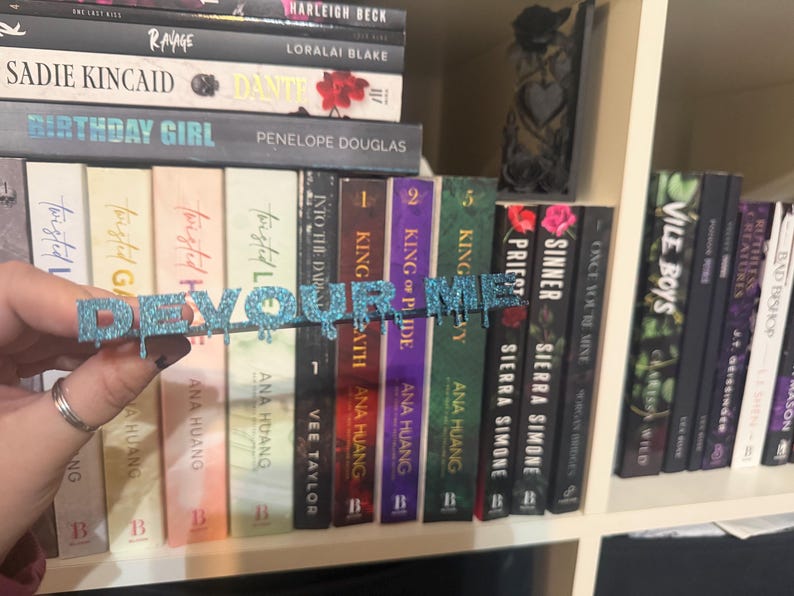 K&ouml;nnte beinhalten: Ein glitzerndes blaues "DEVOUR ME" Buchzubeh&ouml;r wird vor einem B&uuml;cherregal mit B&uuml;chern gehalten. Der Text hat einen tropfenden Effekt. Die B&uuml;cher haben farbenfrohe Buchr&uuml;cken und verschiedene Titel.