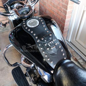Peut inclure: Housse de réservoir de moto en cuir noir avec des clous argentés et une petite pochette avec une boucle. La housse est conçue pour s'adapter aux contours du réservoir de la moto. La moto est noire et chromée.