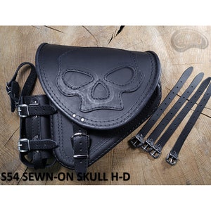 May include: Black leather motorcycle saddlebag with a stitched-on skull design. The bag has a strap with a buckle and a small pouch on the side. The bag is accompanied by four black leather straps with buckles.
