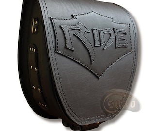 May include: Black leather motorcycle saddlebag with the word "RIDE" embossed on the front. The bag has a shield-shaped design and is stitched with black thread. Gold-colored rivets and hardware are visible on the side.
