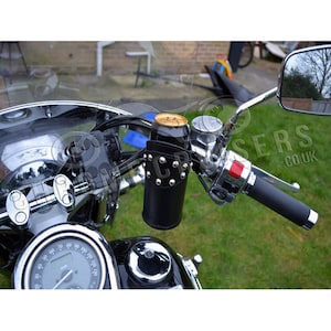 May include: Black leather motorcycle handlebar drink holder with silver studs, holding a gold and tan can. The holder is mounted on a chrome handlebar, with a speedometer and mirror visible. The background shows a residential area.