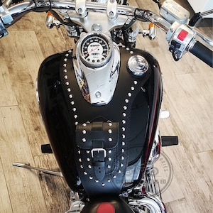 Può includere: Una borsa serbatoio per moto in pelle nera con un design a borchie e una chiusura a fibbia. La borsa è fissata a una moto nera con un serbatoio di benzina cromato e un tachimetro cromato.