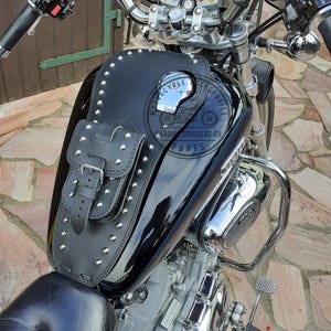 Peut inclure: Bavette de réservoir de moto en cuir noir avec une petite pochette et des clous argentés. La bavette est conçue pour s'adapter à un réservoir de moto et comporte une fermeture à boucle sur la pochette. Le texte "CYCLE ACCESSORIES CHROME CRUISERS EU PARTS" est visible.