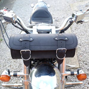 Peut inclure: Une sacoche de selle de moto en cuir noir avec des boucles et des coutures, fixée à un guidon de moto chromé.