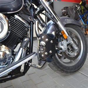 May include: Black leather motorcycle accessory with a silver skull and stud design, mounted on the front fork of a motorcycle. The motorcycle is dark grey with chrome accents. The image also shows a portion of the front tyre and wheel.