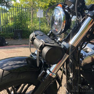 Peut inclure: Gros plan d'une moto noire avec un phare rond et une sacoche à outils en cuir noir fixée à l'avant. La moto présente des détails chromés et un garde-boue noir. L'image est prise à l'extérieur.