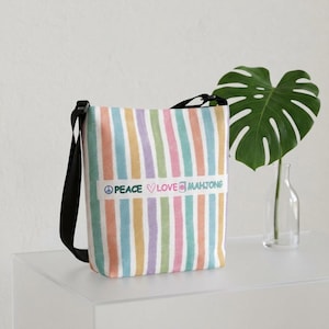 May include: A vertical tote bag featuring a pattern of pastel-colored stripes in pink, blue, yellow, and purple. A white horizontal band across the center displays the text PEACE, a heart symbol, LOVE, a mahjong tile icon, and MAHJONG.