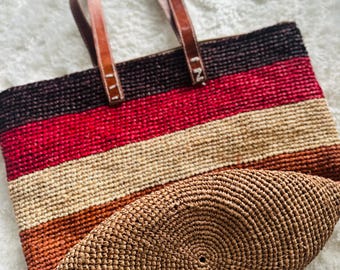 May include: A woven tote bag with brown leather handles. The bag features horizontal stripes in shades of brown, red, and beige. The bottom of the bag has a circular woven design.