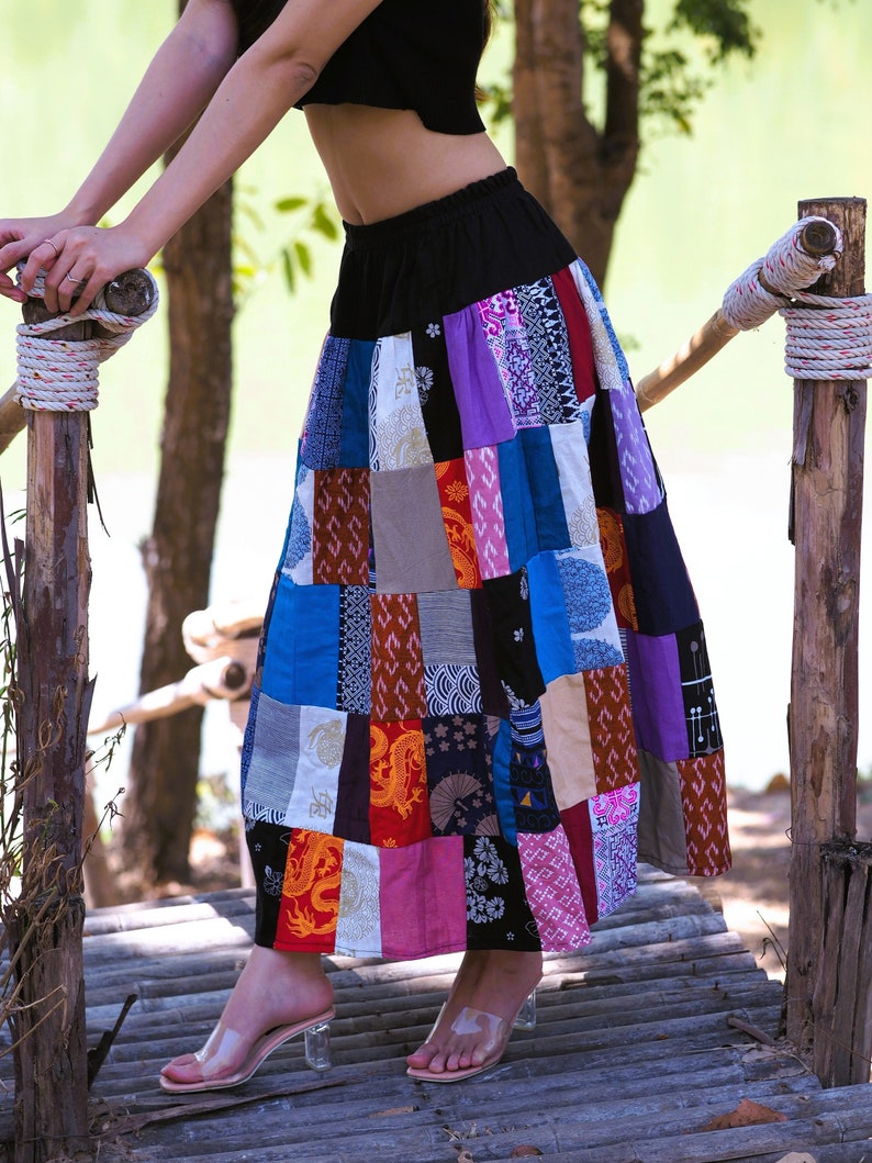 K&ouml;nnte beinhalten: Eine Frau tr&auml;gt einen langen, bunten Patchworkrock mit einem schwarzen Bund. Der Rock besteht aus verschiedenen Stoffen und Mustern, darunter Blumen-, geometrische und Paisley-Muster. Der Rock ist flie&szlig;end und hat eine lockere Passform. Die Frau steht auf einer Holzplattform.