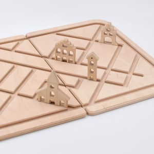 Puede incluir: Un juego de madera con temática de ciudad. El juego incluye una iglesia, una torre, un castillo y un edificio con ventanas. Las piezas están hechas de madera clara y están diseñadas para colocarse en un tablero de madera con un patrón de cuadrícula.