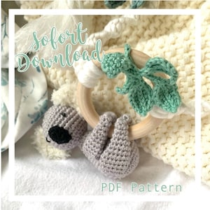Può includere: Un giocattolo koala all'uncinetto appeso a un anello di legno con foglie verdi all'uncinetto. Il giocattolo è su una coperta bianca con il testo "Sofort Download PDF Pattern".