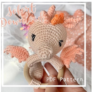 Op de afbeelding: Een gehaakte roze drakenrammelaar met een houten ring. De draak heeft oranje hoorns en zwarte ogen. De tekst "PDF Pattern" is zichtbaar in de afbeelding.