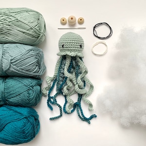 Op de afbeelding: Een gehaakte kwallen speelgoedset met groen en blauw garen, vulling, houten kralen en touw.