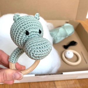 Puede incluir: Un sonajero de hipopótamo de crochet de color verde claro con un anillo de madera. El sonajero está en una caja con otros artículos para bebés, incluyendo un cuadrado de tela verde y un anillo de madera.