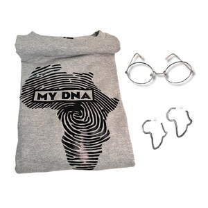 May include: A gray t-shirt with a black print of the continent of Africa in the shape of a fingerprint. The text "MY DNA" is printed in a white rectangle on the print. The t-shirt is folded and laid flat on a white background. There are also silver round glasses and silver earrings shaped like the continent of Africa.