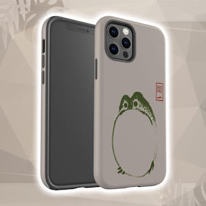 May include: A smartphone with a light beige case featuring a green and black illustration of a frog-like creature inside a circle. The case also has a red and black Japanese character. The phone is displayed next to a black smartphone.