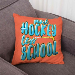 Könnte beinhalten: Ein orangefarbenes quadratisches Kissen mit dem Text "more HOCKEY less SCHOOL" in einer blau-weißen Blasen-Schriftart. Eine gelb-braune Hockeyschläger-Grafik befindet sich zwischen den Wörtern. Das Kissen liegt auf einem grauen Sofa.