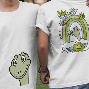 May include: Two white t-shirts with a green dinosaur graphic. One shirt features a dinosaur head peeking out from behind a rainbow and flowers. The other shirt features a full dinosaur head.