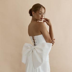 Op de afbeelding: Een witte strapless bruidsjurk met een grote strik op de rug. De jurk heeft een getailleerd lijfje en een zwierige rok.