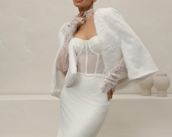 May include: A white bridal ensemble featuring a fitted lace gown, a sheer corset top, and a fluffy, short-sleeved jacket. The look is completed with delicate lace gloves. The outfit is perfect for a wedding or special occasion.